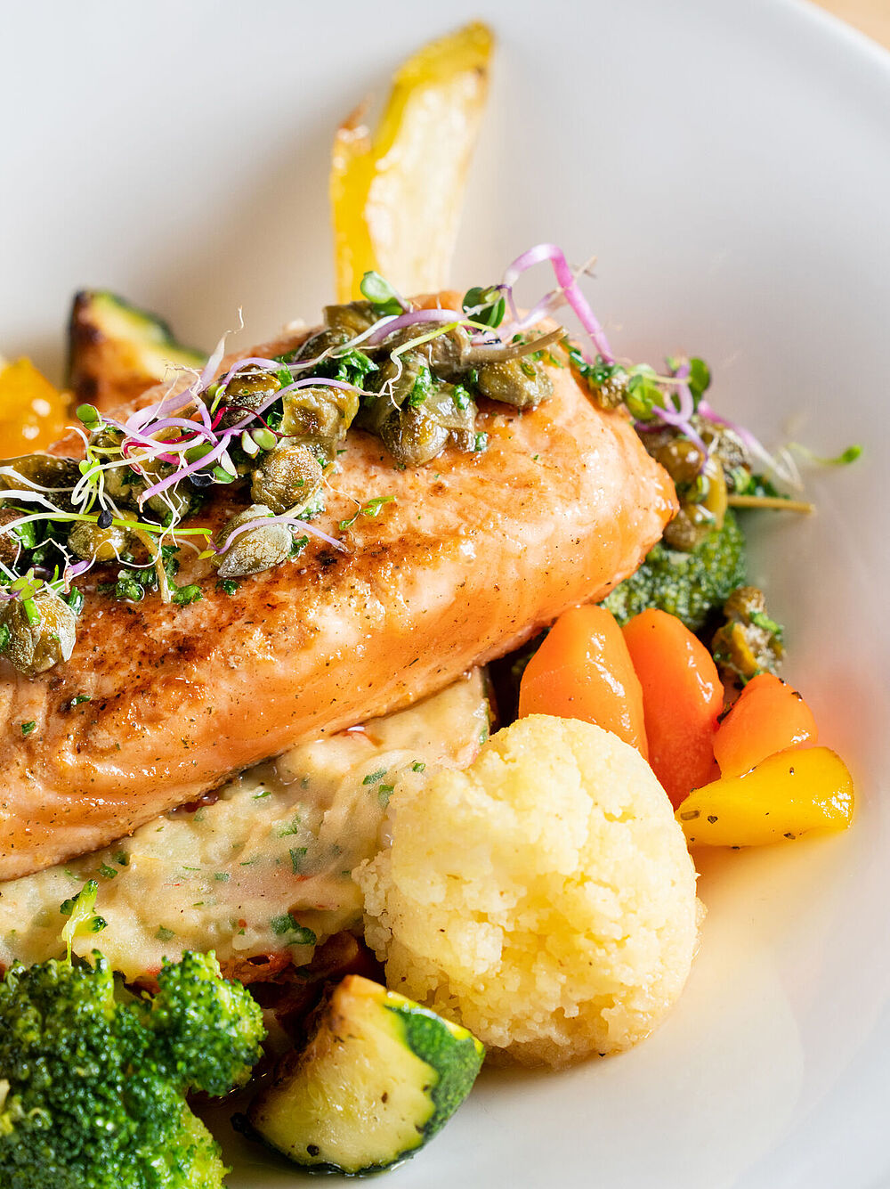 Grilled salmon fillet on puree, garnished with herbs, served with colourful vegetables: broccoli, carrots and yellow peppers on a white plate.