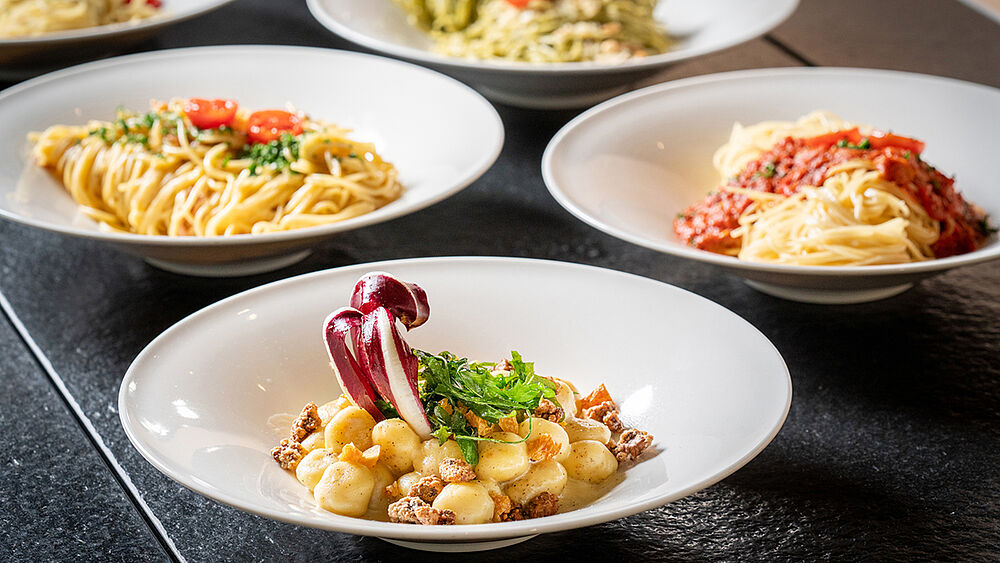 Eine Vielzahl von Nudelgerichten auf einem dunklen Tisch. Auf jedem weißen Teller liegen verschiedene Nudeln mit Soßen und Beilagen, die ein Gefühl von köstlicher italienischer Küche vermitteln.