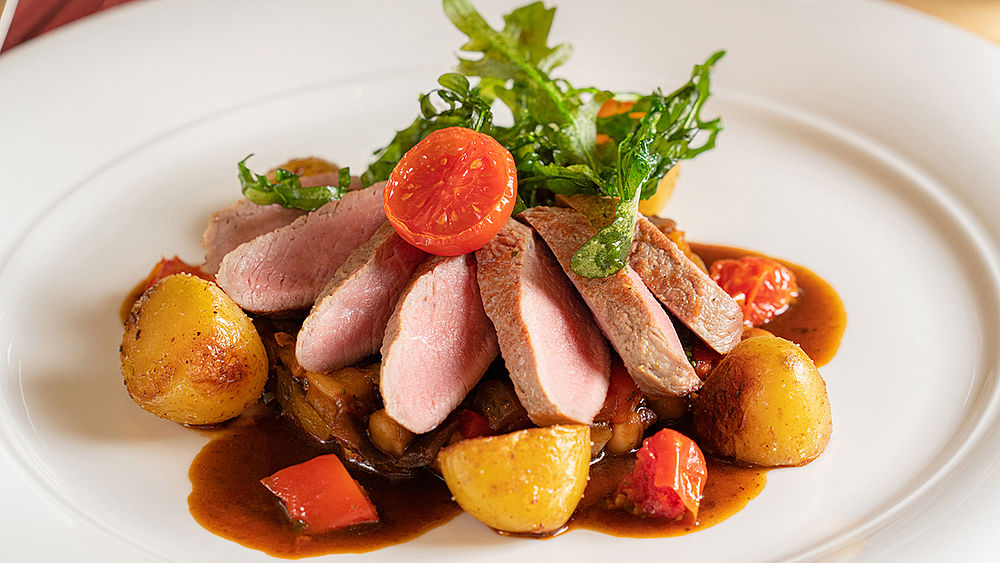 Gourmet-Gericht mit Entenbrustscheiben auf Röstkartoffeln, Kirschtomaten und Grünzeug, beträufelt mit reichhaltiger brauner Sauce auf einem weißen Teller.
