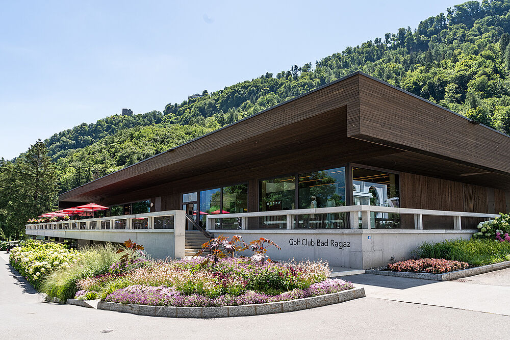 Modernes Gebäude mit einem scharfen, kantigen Flachdach vor einem Wald und einer Bergkulisse. Die sonnige Beleuchtung schafft ein warmes, einladendes Ambiente.