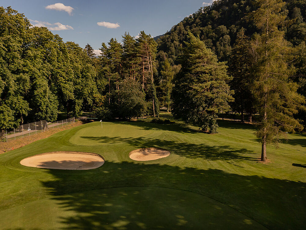 Ein Golfplatz mit zwei Sandbunkern ist von Bäumen unter einem strahlend blauen Himmel umgeben. Die Schatten sorgen für eine ruhige und friedliche Atmosphäre.
