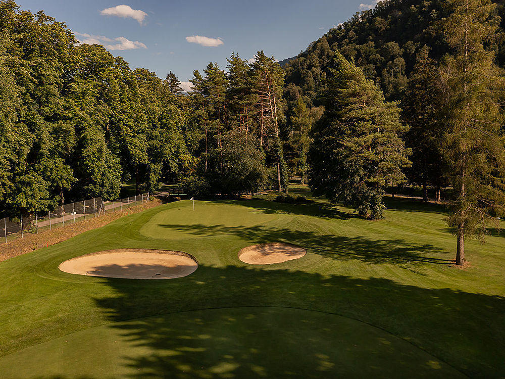 A golf course with two sand bunkers is surrounded by trees under a bright blue sky. The shadows create a calm and peaceful atmosphere.