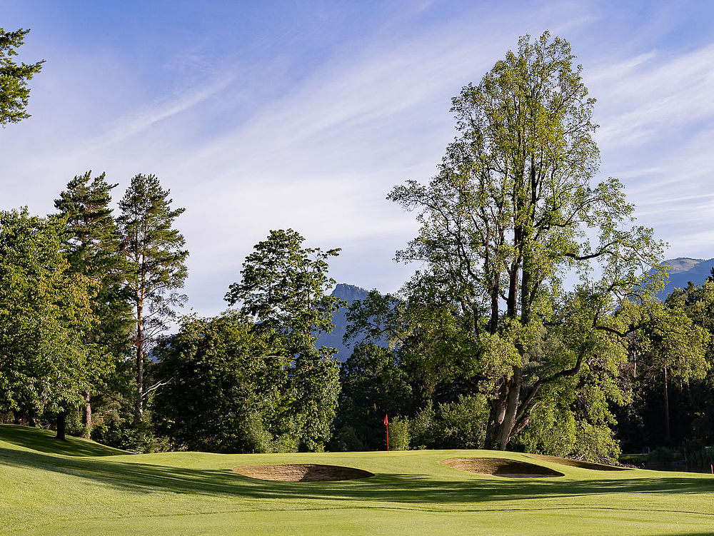 The golf course at hole 14 with tall trees under a blue sky. The sunlight falls through the leaves and creates a calm and peaceful atmosphere. Two bunkers in the foreground and Lake Giessen to the right of the picture.