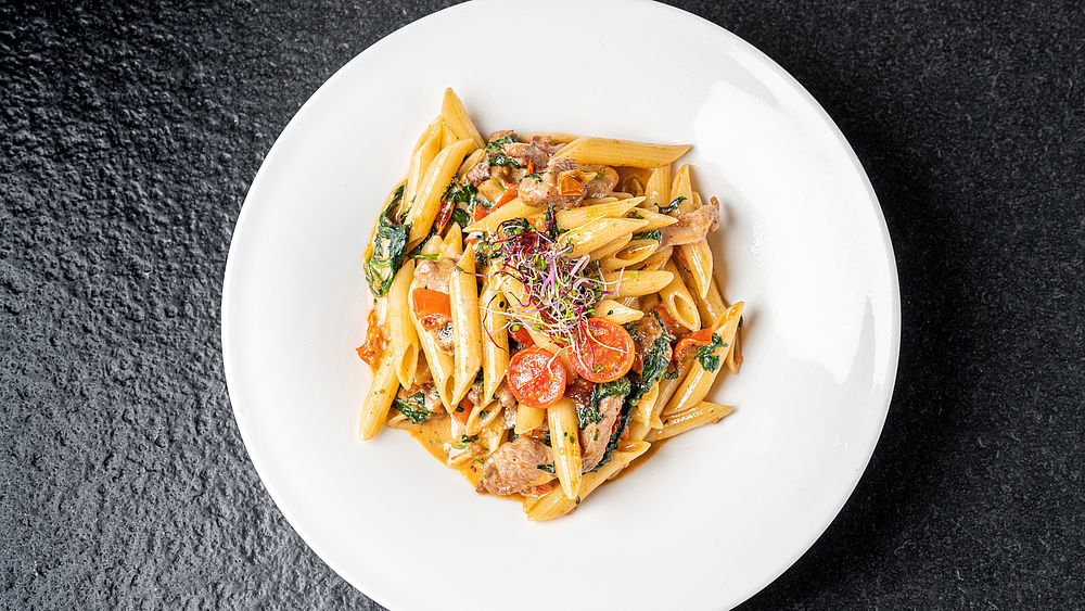 Auf einem weissen Teller mit dunkler Oberfläche liegen Penne mit cremiger Sauce, Kirschtomaten und Kräutern, die eine farbenfrohe und appetitliche Präsentation ergeben.