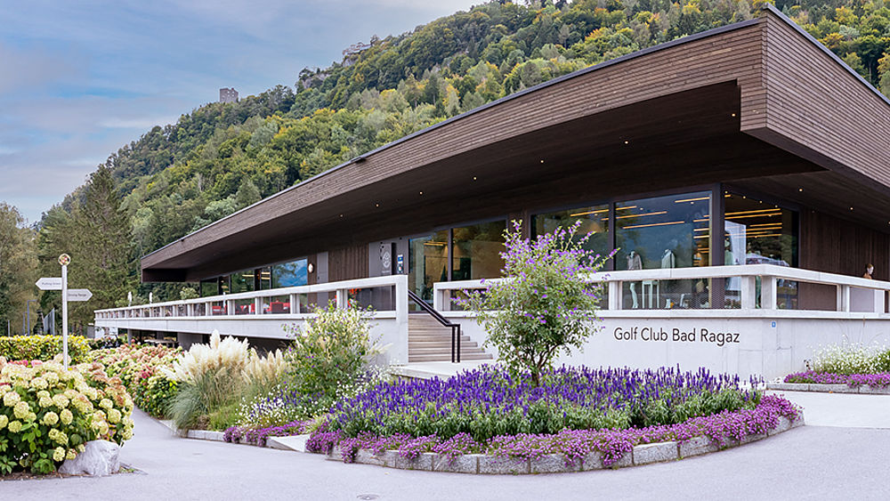 The Bad Ragaz clubhouse is a modern wooden building with large glass windows, surrounded by colourful flowerbeds with purple and white blossoms and set in the middle of a green hill.