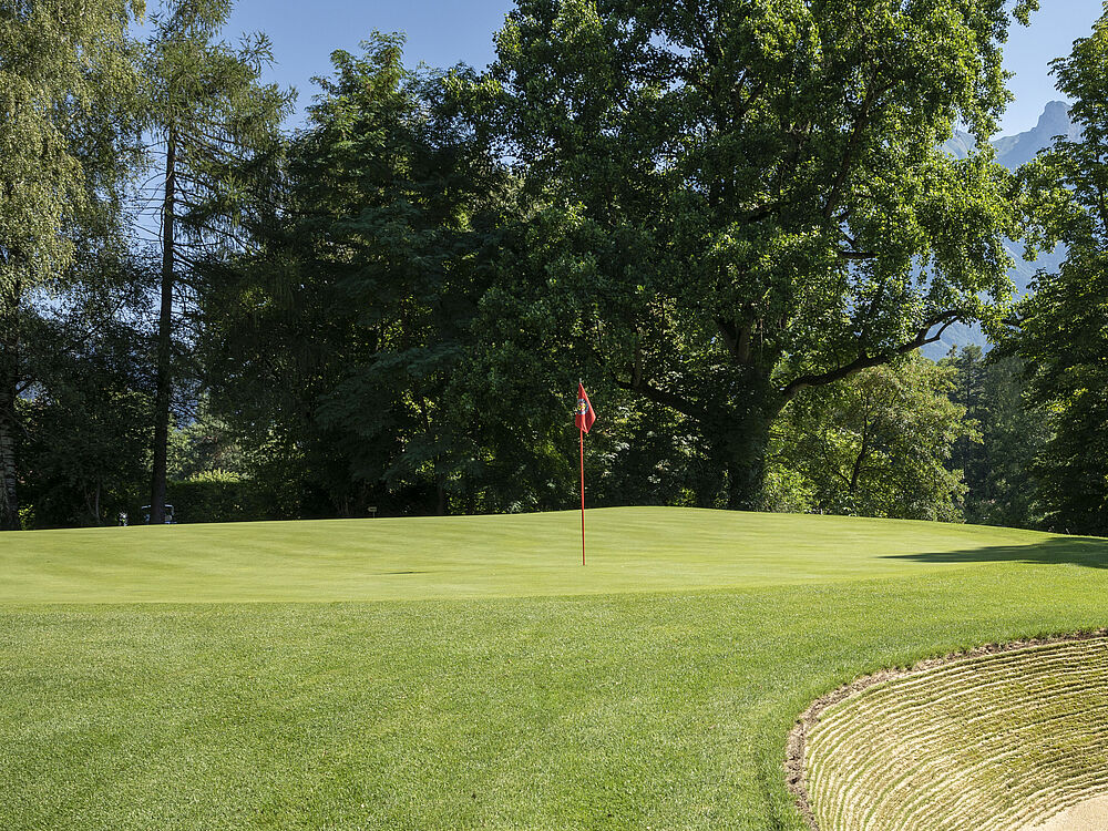 Ein gepflegter Golfplatz mit üppigem grünem Gras, einem Sandbunker auf der rechten Seite und einer roten Flagge, die das Loch markiert. Im Hintergrund hohe Bäume und Berge.