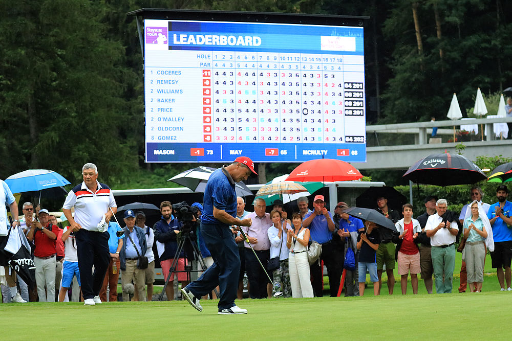 Ein Golfer in Blau jubelt auf dem Grün mit einem Fist Pump, umgeben von Zuschauern mit Regenschirmen. Im Hintergrund ist eine Anzeigetafel mit den Spielergebnissen zu sehen.