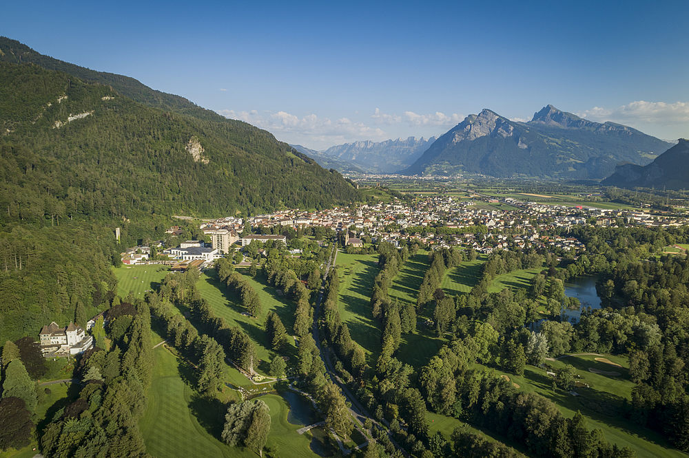 Luftaufnahme eines Golfplatzes mit sattgrünen Fairways, umgeben von dichten Wäldern und Bad Ragaz, vor dem Hintergrund der fernen Berge.