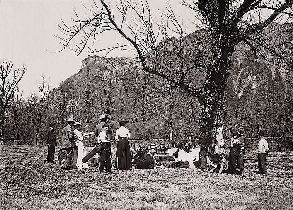 Vintage-Foto einer Gruppe, die ein Picknick unter einem Baum in einer bergigen Gegend genießt. Männer, Frauen und Kinder entspannen sich; die Szene wirkt ruhig und nostalgisch.