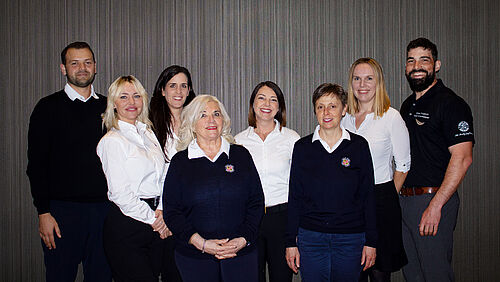8 employees next to each other. Dressed in dark jumpers and white shirts against a dark background.