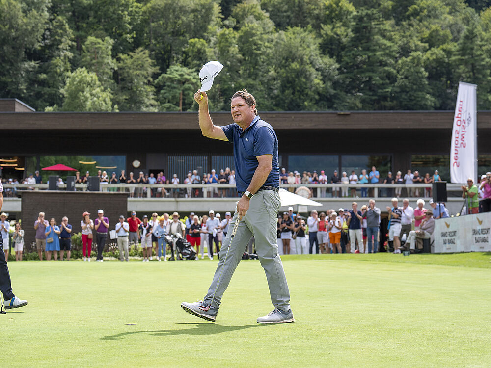 Ein Golfer auf dem Grün winkt der jubelnden Menge mit seiner Mütze zu. Er trägt ein blaues Hemd und eine graue Hose. Im Hintergrund sind Bäume und ein großes Clubhaus zu sehen.