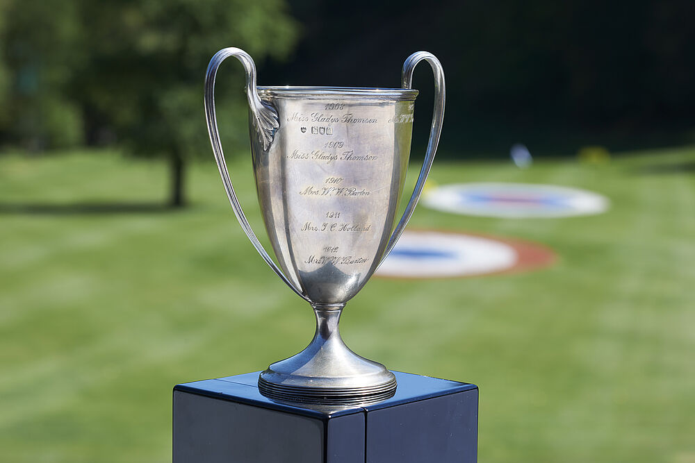 Silberne Trophäe mit geschwungenen Griffen auf einem Sockel vor dem Hintergrund des grünen Golfplatzes. Hell und sonnig, ein Gefühl des Sieges und der Leistung hervorrufend.