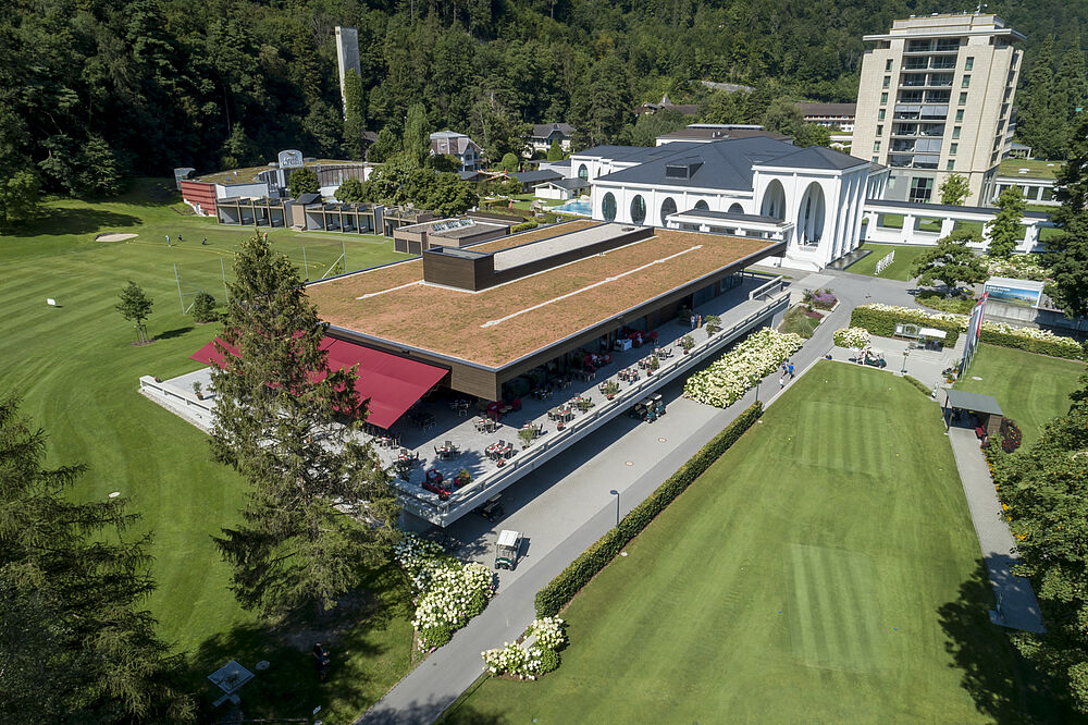 Luftaufnahme eines grünen Golfplatzes mit einem modernen Clubhaus und einem hohen Gebäude im Hintergrund, vor einem bewaldeten Hügel. Helle und heitere Atmosphäre.