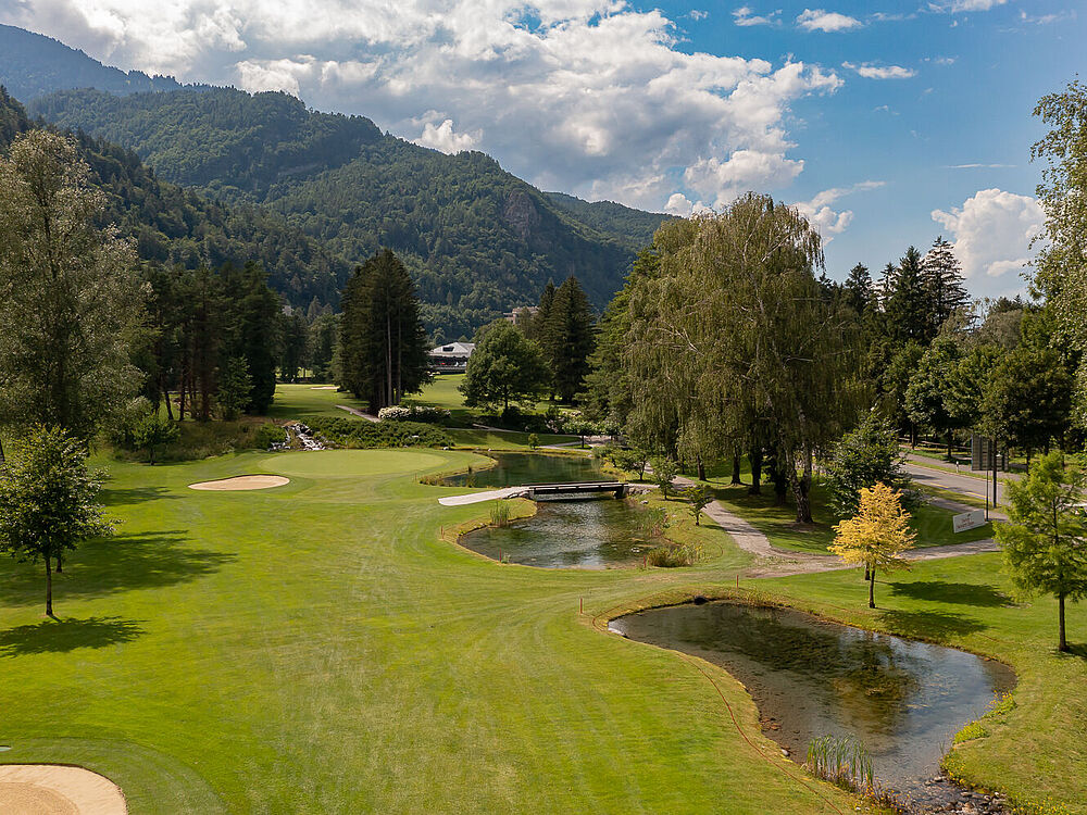 Ein Golfplatz mit grünen Fairways, einem kleinen gewundenen Bach und Bäumen unter einem teilweise bewölkten Himmel. Die Berge bilden eine malerische Kulisse. Friedliche Umgebung.