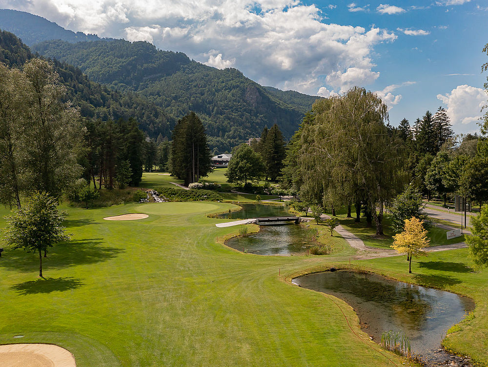 A golf course with green fairways, a small winding stream and trees under a partly cloudy sky. The mountains form a picturesque backdrop. Peaceful surroundings.