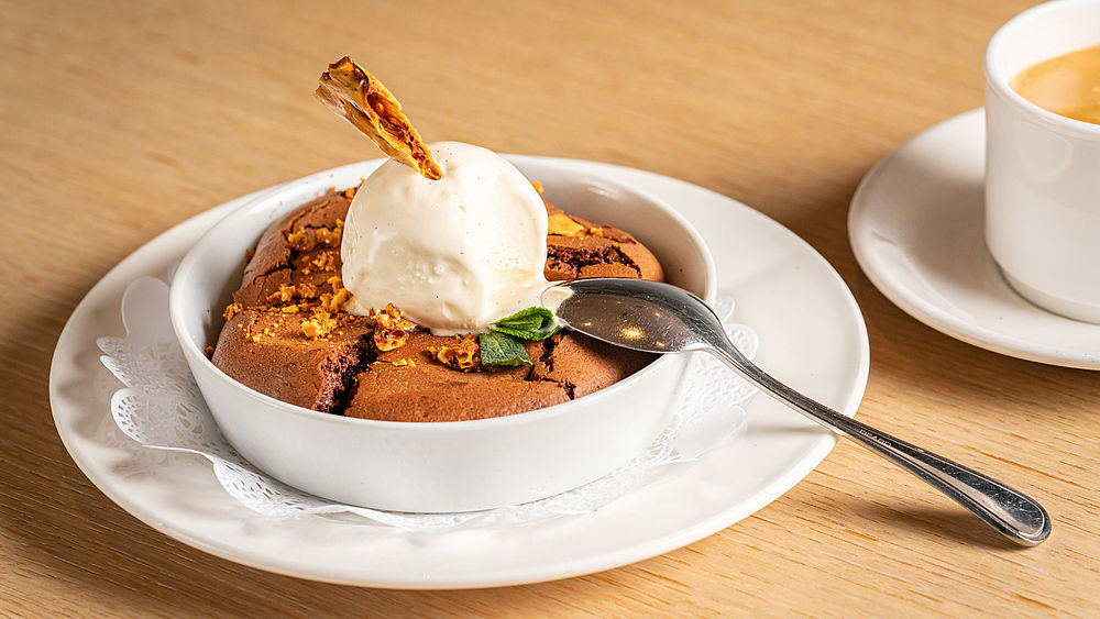 Ein warmer Brownie in einer weissen Schale, garniert mit einer Kugel Vanilleeis und Karamellstückchen, garniert mit Minze. Ein Löffel liegt daneben, eine Tasse Kaffee steht im Hintergrund.