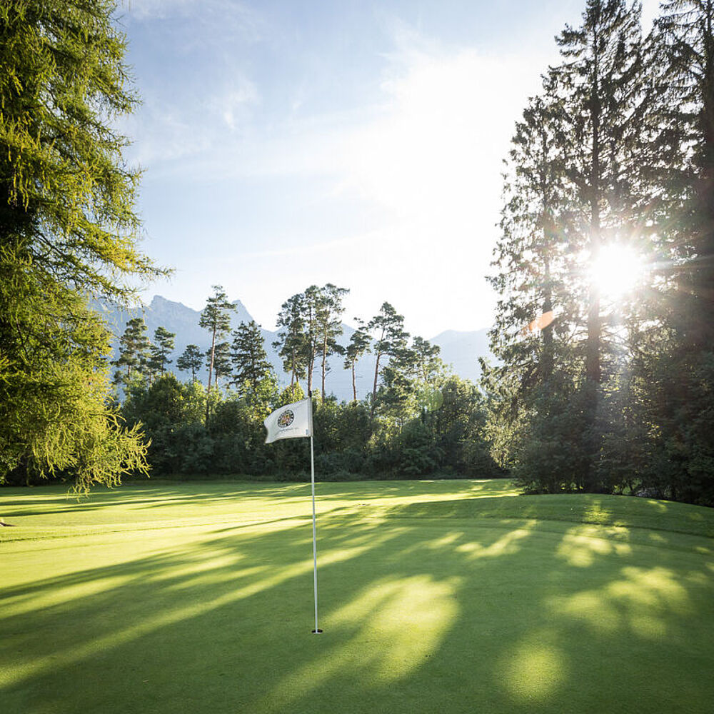 Ein sonniger Golfplatz mit einer Fahne und einem Loch im Vordergrund. Im Hintergrund sind hohe Bäume und Berge, die lange Schatten werfen. Friedliche Atmosphäre.