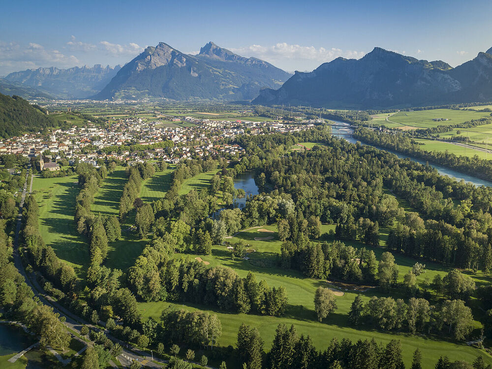 Luftaufnahme des Golfplatzes mit Wäldern, einem sich schlängelnden Fluss und einer kleinen Stadt, die sich unter einem klaren blauen Himmel an die Berge schmiegt.