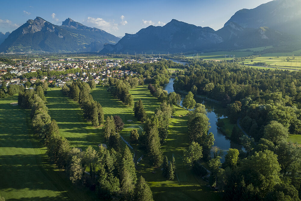 Luftaufnahme des Golfplatzes mit einem gewundenen Fluss, der von dichten Bäumen gesäumt wird. Eine kleine Stadt schmiegt sich an den Fuß der majestätischen Berge unter einem klaren blauen Himmel.