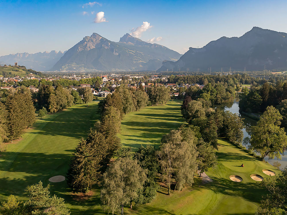 A green golf course with tall trees and a small pond against the backdrop of majestic mountains under a bright blue sky, creating a serene atmosphere.