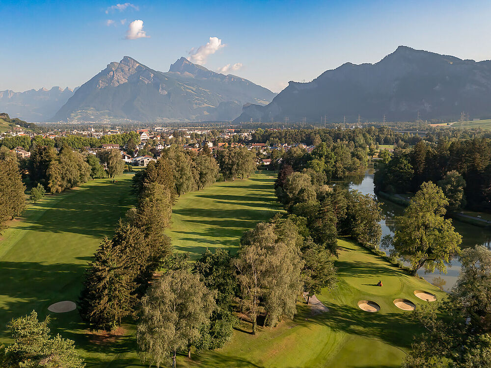 Ein grüner Golfplatz mit hohen Bäumen und einem kleinen Teich vor der Kulisse majestätischer Berge unter einem strahlend blauen Himmel, der eine heitere Atmosphäre schafft.