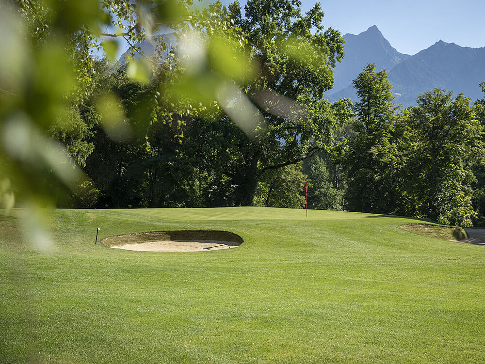 Ein grüner Golfplatz mit Sandbunkern, umgeben von Bäumen und vor der Kulisse der fernen Berge unter einem klaren blauen Himmel.