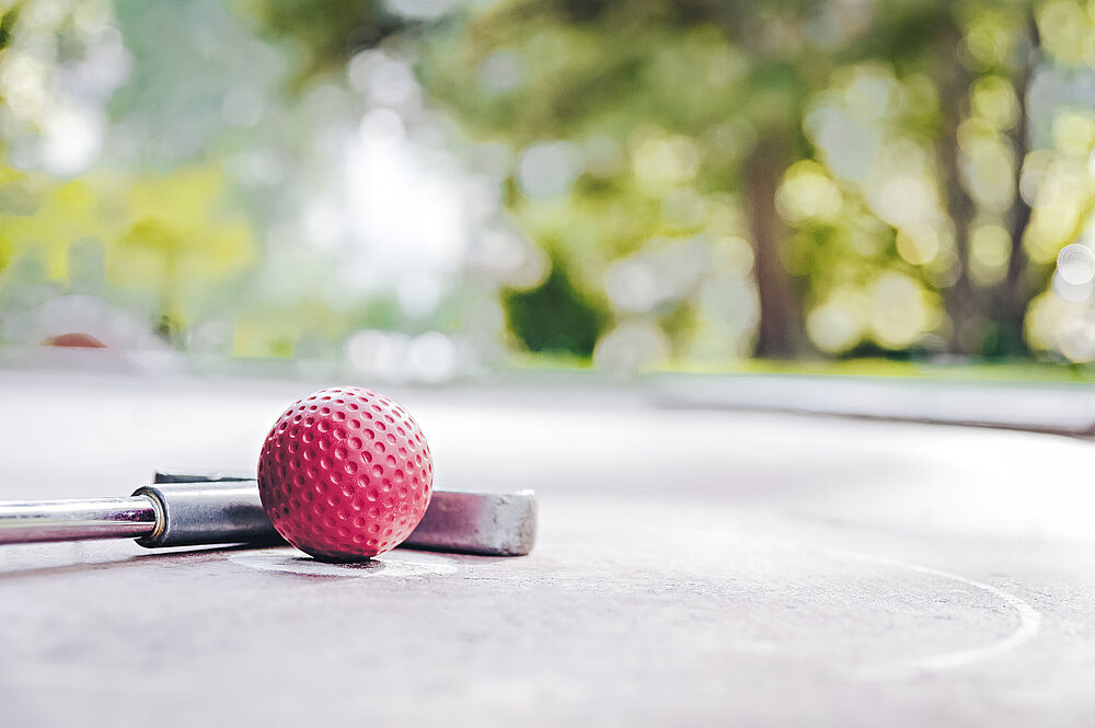 Ein Minigolfschläger liegt am Boden im Vordergrund ein Golfball in rot. Im Hintergrund sieht man den Wald.