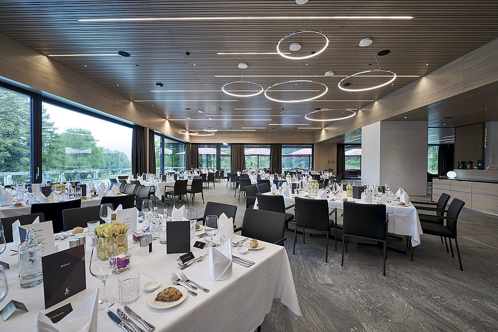 Modernes Restaurant mit grossen Fenstern, elegant gedeckten Tischen mit weissen Tischtüchern und schwarzen Stühlen. Sanfte Deckenbeleuchtung schafft ein anspruchsvolles Ambiente.