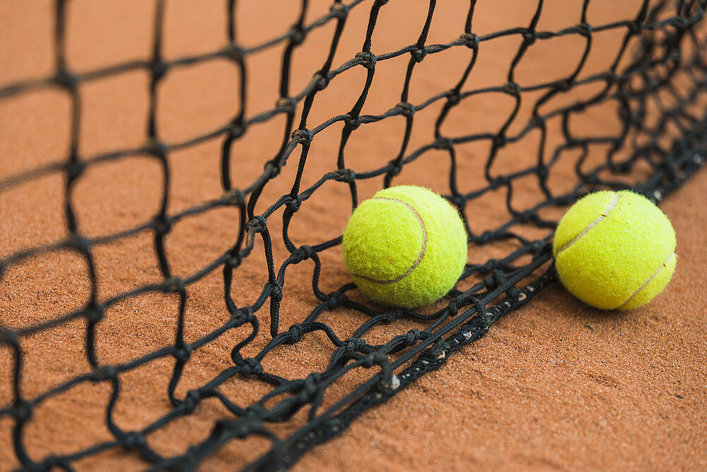 Zwei Tennisbälle, die auf einem Sandplatz vor einem schwarzen Netz ruhen. Die Szene vermittelt einen ruhigen, innegehaltenen Moment bei einem Tennisspiel und betont Sport und Freizeit.