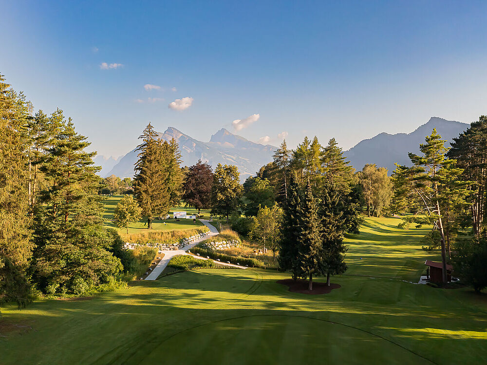 Ein malerischer Golfplatz, umgeben von hohen Bäumen und Bergen unter einem klaren blauen Himmel. Das Sonnenlicht wirft sanfte Schatten und schafft eine ruhige und einladende Atmosphäre.
