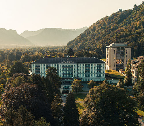 Luftaufnahme des Grand Resort Bad Ragaz, umgeben von üppigen grünen Wäldern und Hügeln bei Sonnenuntergang. Die elegante Architektur des Anwesens wird durch das warme goldene Licht hervorgehoben.