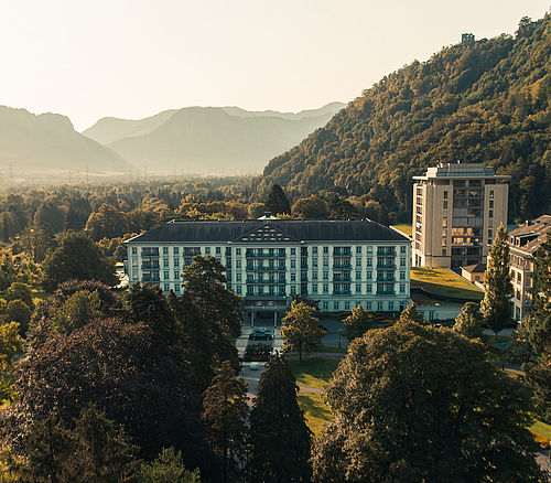 Aerial view of the Grand Resort Bad Ragaz, surrounded by lush green forests and hills at sunset. The elegant architecture of the property is emphasised by the warm golden light.