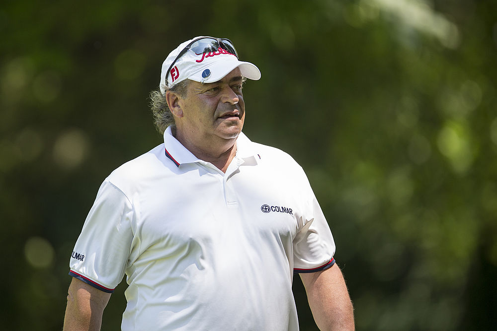 Ein Mann in einem weißen Polohemd und einer Kappe steht auf einem Golfplatz und schaut konzentriert. Der Hintergrund ist üppig und grün und vermittelt einen ruhigen, sonnigen Tag.