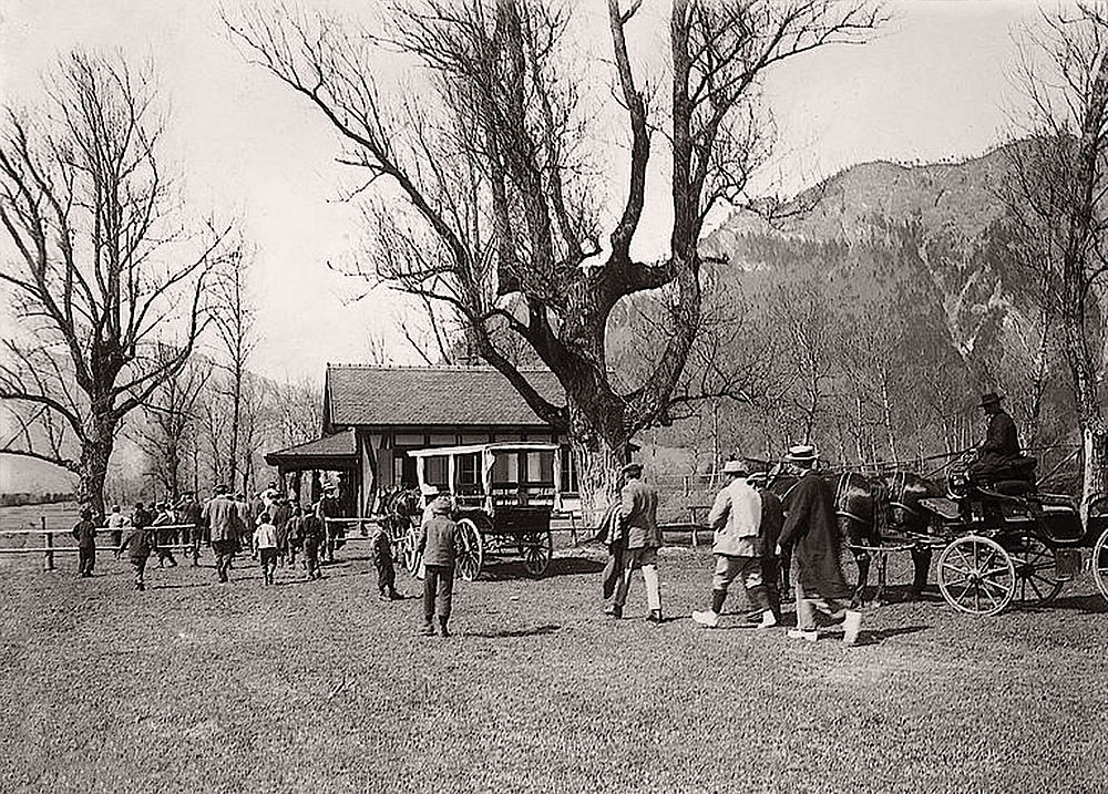 Vintage-Foto eines Treffens mit Freunden im Freien, mit Menschen, die sich in der Nähe eines Hauses und von Bäumen treffen. Die Szene vermittelt Gemeinschaft und Wärme in einer ländlichen Umgebung.