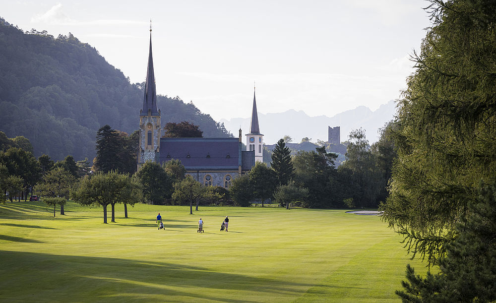 Ein ruhiger Golfplatz mit Spielern im Vordergrund, umgeben von üppigem Grün. Eine Kirche mit zwei Türmen vor dem Hintergrund nebliger Berge.
