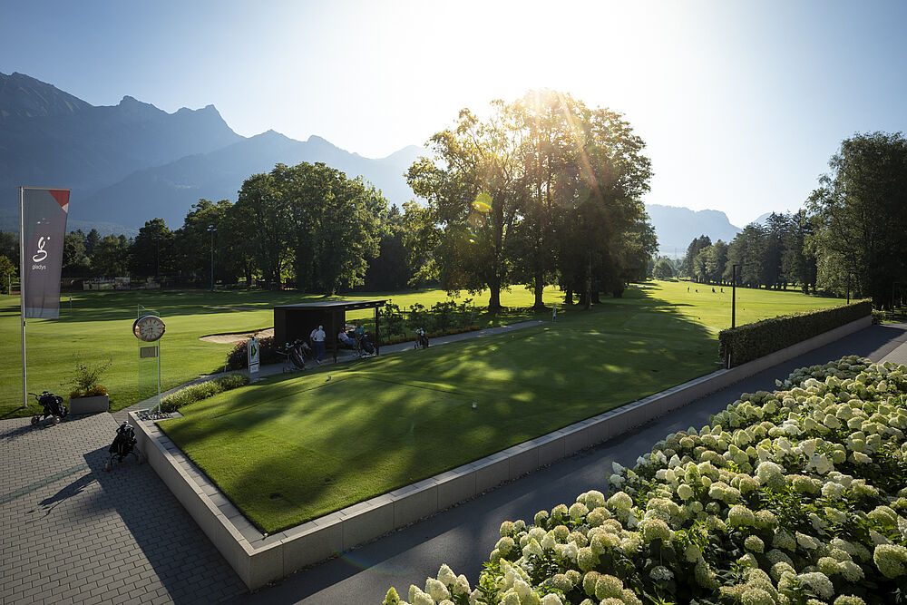Sonnenbeschienener Park mit üppigem grünen Gras und verstreuten Bäumen, Berge im Hintergrund. Ein kleiner Pavillon und eine Fahne sind in der Szene zu sehen, die Ruhe ausstrahlen.