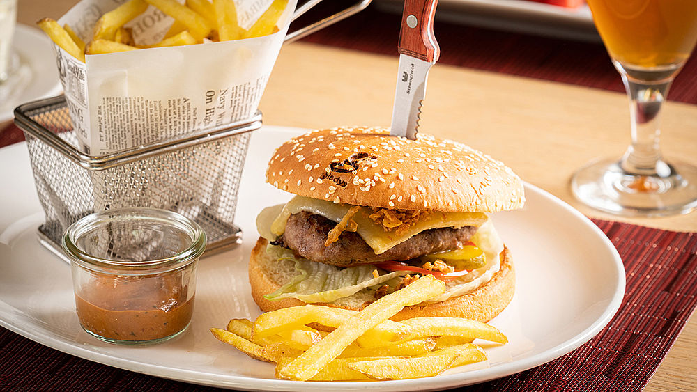 Ein klassischer Cheeseburger mit Salat und Pommes frites auf einem weissen Teller, begleitet von einem kleinen Drahtkorb mit Pommes frites und einem Getränk, auf einem Holztisch.