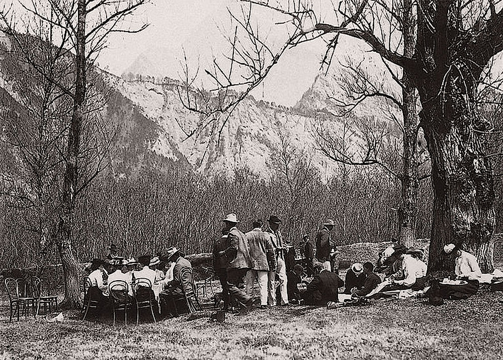 Eine Gruppe Menschen aus 1900 sitzt und steht unter Bäumen vor der Kulisse der hoch aufragenden Berge. Die Szene erinnert an eine heitere, historische Atmosphäre.