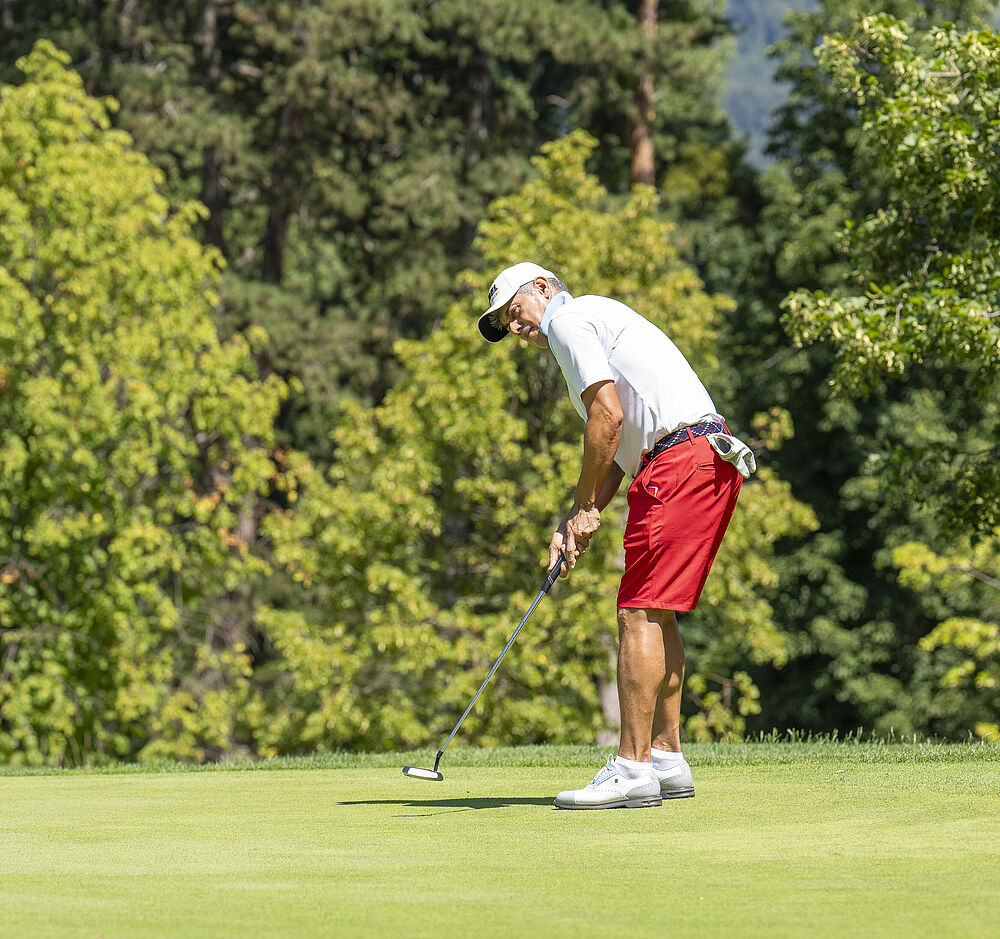 Ein Golfer in weißem Hemd und roten Shorts konzentriert sich beim Putten auf einem üppig grünen Platz, umgeben von leuchtenden Bäumen unter klarem Himmel.
