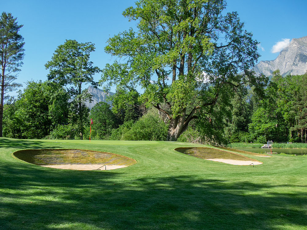 Landschaftlich reizvoller Golfplatz mit zwei Sandbunkern auf einem üppig grünen Fairway, umgeben von hohen Bäumen und fernen Bergen unter einem strahlend blauen Himmel.