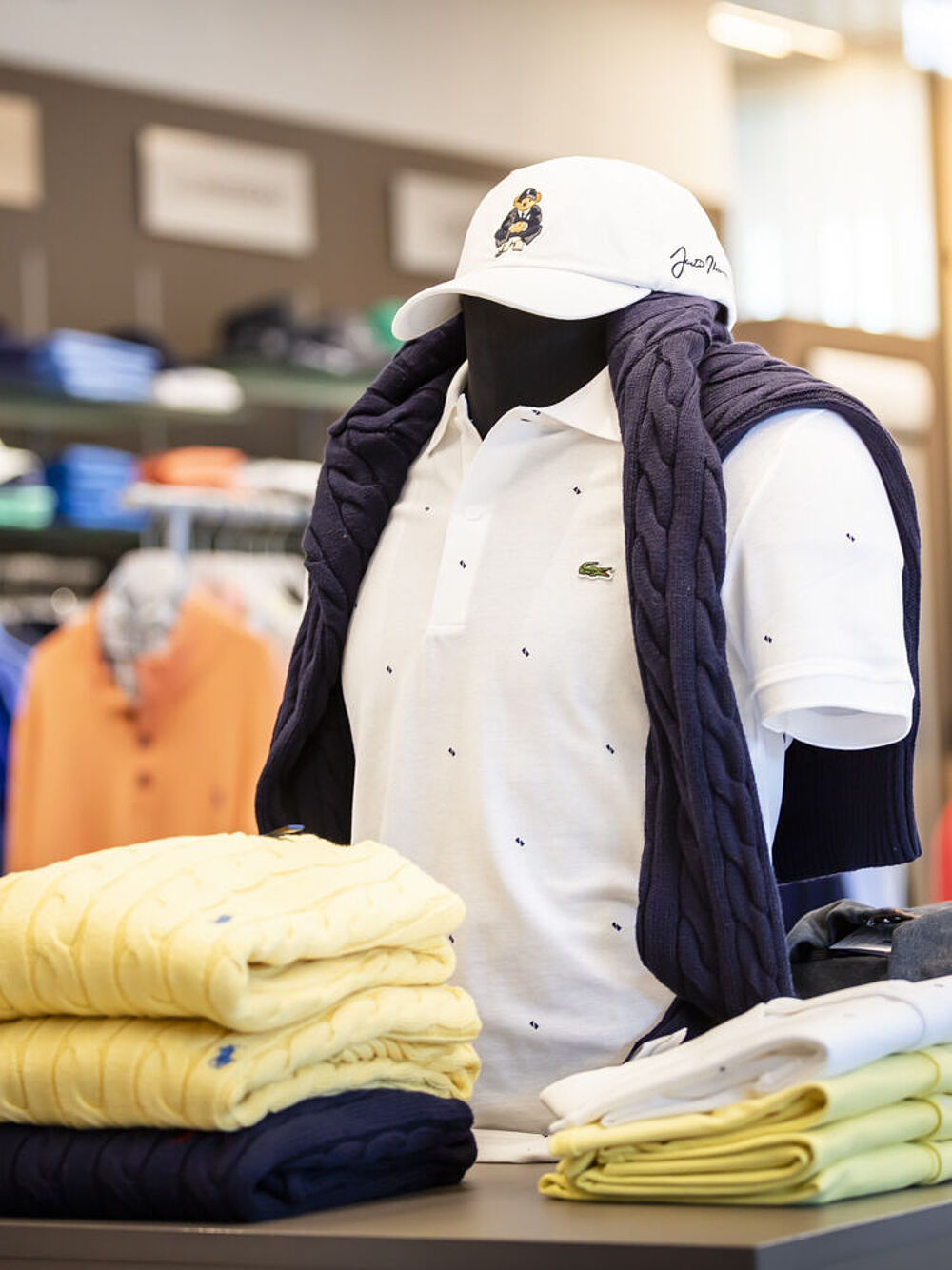 Mannequin in golf shop wearing a white polo shirt and cap with a navy blue jumper pulled over her shoulders. Yellow and white shirts folded nearby. Casual and stylish atmosphere.