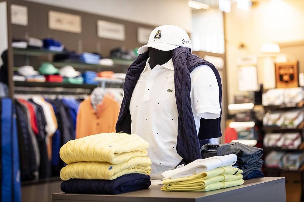 Schaufensterpuppe im Golf Shop, die ein weißes Polohemd und eine Mütze trägt und einen marineblauen Pullover über die Schultern gezogen hat. Gelbe und weiße Hemden in der Nähe gefaltet. Lässige und stilvolle Atmosphäre.