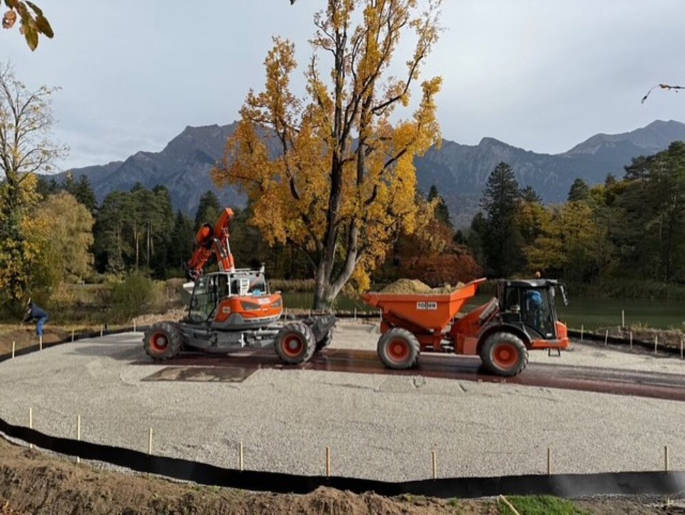 Baufahrzeuge arbeiten auf einem Schotteruntergrund in der Nähe eines großen Herbstbaums mit Bergen im Hintergrund und vermitteln ein Gefühl von Industrie inmitten der Natur.