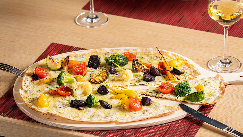 Eine bunter Flammkuchen, belegt mit Kirschtomaten, Oliven und Kräutern auf einem Holzbrett. Im Hintergrund stehen zwei Gläser mit Weißwein.