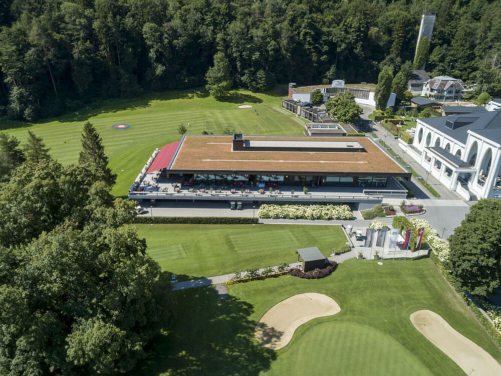 Luftaufnahme eines modernen Golfclubhauses mit grünem Dach, umgeben von üppigen Bäumen und einem gepflegten Golfplatz. Die Umgebung ist ruhig und einladend.
