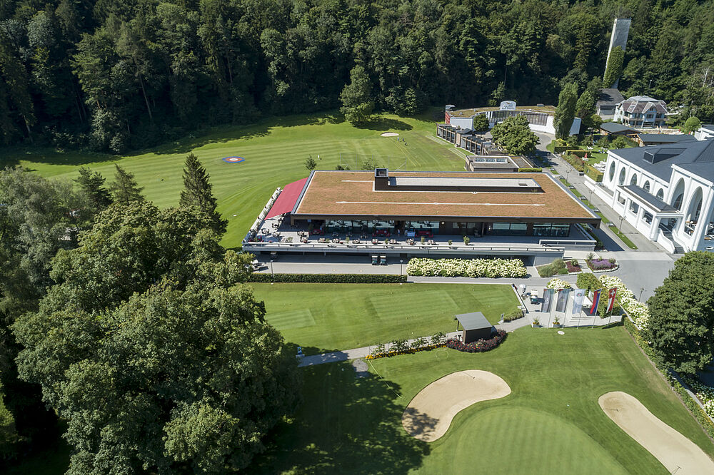 Luftaufnahme eines modernen Golfclubhauses mit grünem Dach, umgeben von üppigen Bäumen und einem gepflegten Golfplatz. Die Umgebung ist ruhig und einladend.