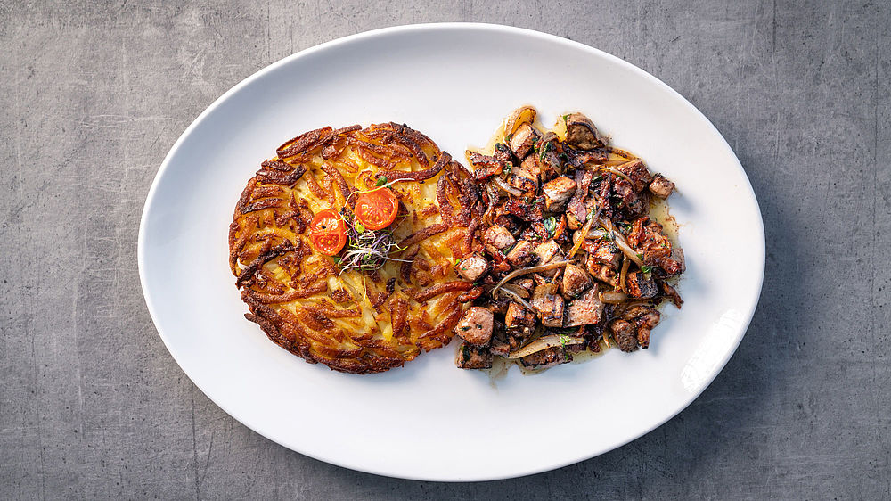 On a white plate lies a golden-brown rösti, garnished with cherry tomatoes and herbs, next to a portion of fried mushrooms and sliced liver, creating a rustic, appetising presentation.