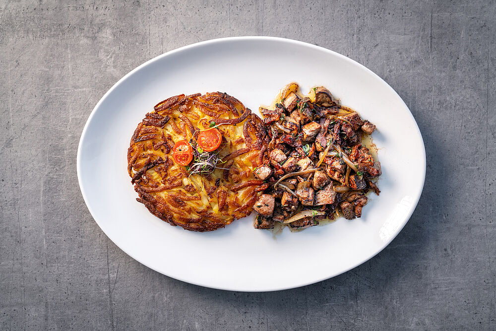 Auf einem weissen Teller liegt ein goldbrauner Rösti, garniert mit Kirschtomaten und Kräutern, neben einer Portion gebratener Pilze und Lebergeschnetzeltem, die eine rustikale, appetitliche Präsentation ergeben.