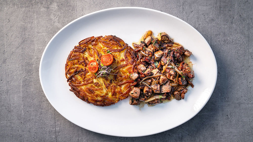 Auf einem weissen Teller liegt ein goldbrauner Rösti, garniert mit Kirschtomaten und Kräutern, neben einer Portion gebratener Pilze und Lebergeschnetzeltem, die eine rustikale, appetitliche Präsentation ergeben.