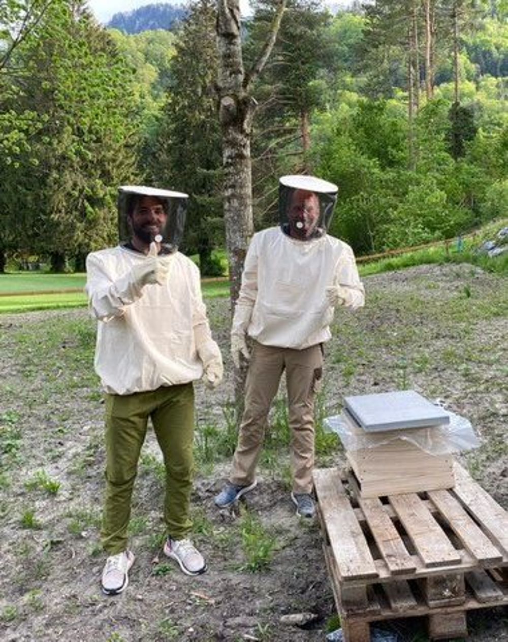Zwei Personen in Imkeranzügen stehen im Freien neben Bienenstöcken auf Holzpaletten. Sie sind von üppigem Grün umgeben und scheinen zufrieden zu sein.